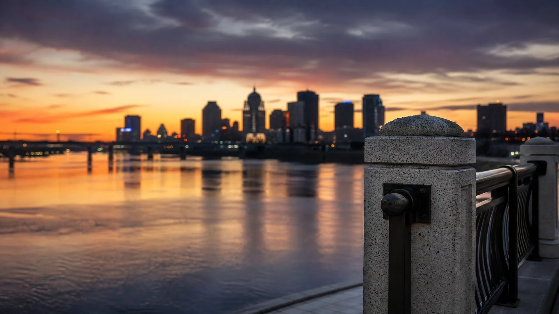 A city skyline silhouetted against an orange and purple sunset sky, reflected in a wide river, viewed past a blurred stone post and metal railing in the foreground.